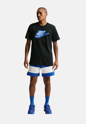 Hombre con camiseta negra "Just Do It", pantalones cortos blancos y azules, calcetines azules y zapatillas azules de pie frente a un fondo blanco.
