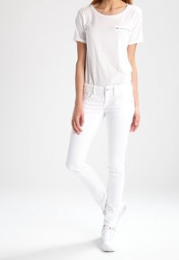 Camiseta blanca de manga corta con bolsillo, combinada con jeans blancos ajustados y zapatillas blancas. Tejido suave y líneas limpias en toda la prenda.