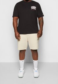 Camiseta negra de manga corta con el logo "TOMMY", combinada con pantalones cortos beige claro. Calcetines deportivos blancos y zapatillas completan el conjunto.