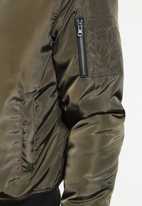 Bomber bunda olivově zelená s lesklou hladkou texturou. Má kapsu na zip a kontrastní černé manžety a doplňky.