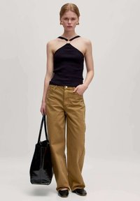 Zwarte geribbelde tanktop met halternek, gecombineerd met losse bruine broek en een zwarte glanzende totebag. Platte zwarte schoenen maken de look af.