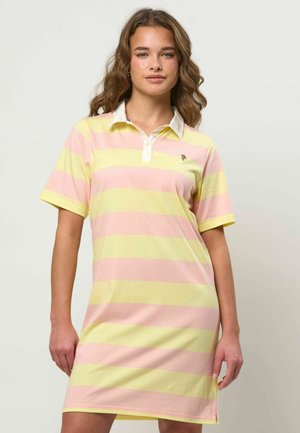 Femeie purtând o rochie polo cu mâneci scurte, cu dungi late orizontale roz și galbene, stând în picioare pe un fundal simplu, deschis.