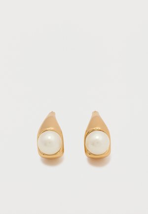 Σκουλαρίκια καρφάκια σε χρυσό τόνο, το καθένα με μία στρογγυλή λευκή πέρλα, τοποθετημένη σε λείο, κυρτό μέταλλο σε απλό φωτεινό φόντο.