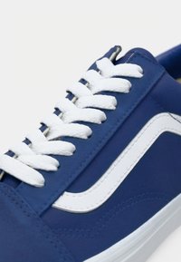 Blaue Canvas-Sneaker mit einem weißen Seitenstreifen, weißen Schnürsenkeln und Nähten. Das Design umfasst eine abgerundete Zehenpartie und eine flache Sohle.