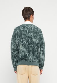 Mīksts, zaļš teksturēts džemperis ar noslēpumainu rakstu un ribbētu apakšmalu, apvienots ar gaišām bezkrāsām biksēm. Redzama šuvju detaļa uz biksēm.