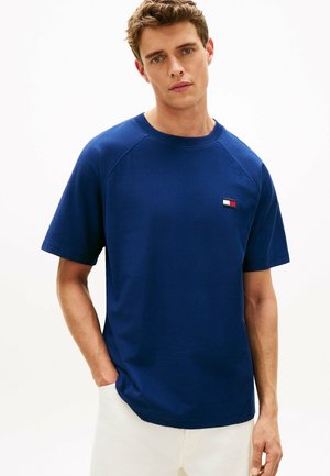 Junger Mann, der ein blaues Kurzarmhemd mit einem kleinen rechteckigen Logo trägt, mit einer Hand in der Tasche, vor einem schlichten weißen Hintergrund stehend.