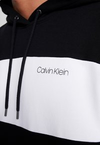 Felpa con cappuccio bianco e nero con una striscia orizzontale bianca e il logo "Calvin Klein" in carattere nero. Tessuto strutturato, cordini regolabili.