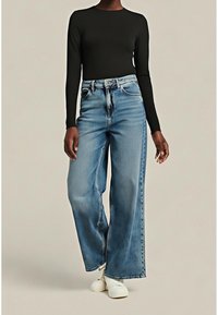 Personne portant un haut noir à manches longues, un jean bleu taille haute à jambes larges, et des baskets blanches, debout devant un fond beige uni.
