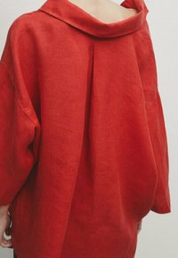 Chemise rouge en lin avec un col large, une coupe décontractée et des épaules tombantes. Elle présente un pli à l'arrière pour plus de détail.