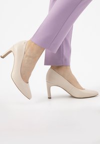 Scarpe col tacco beige con punta affusolata e finitura in pelle liscia, appoggiate su una superficie di colore chiaro, abbinate a pantaloni viola.