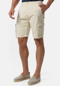 Man in beige cargo shorts met zijvakken en lichtgrijze instap espadrilles, staand tegen een witte achtergrond.