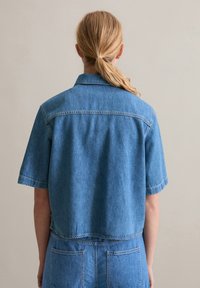 Short-sleeved denim shirt in light blue, with a collar, back yoke seam, and a cropped design. Pockets visible at the waist.