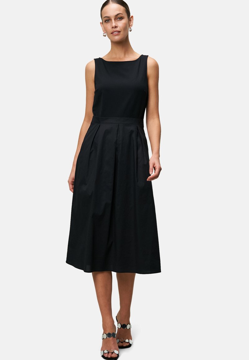 Black sleeveless dress with a boat neckline, fitted bodice, and pleated skirt. Smooth fabric with a mid-calf length. Accented with black sandals.