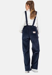 Marine blauw corduroy overall met verstelbare schouderbanden, wijde pijpen en twee achterzakken. Bevat een decoratieve patch op de rechterzak.
