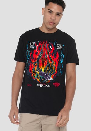 Man wearing black Stranger Things t-shirt with red, blue, and orange graphic of a figure surrounded by flames and spider-like legs.