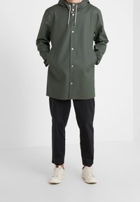 Mężczyzna w ciemnozielonym płaszczu przeciwdeszczowym z kapturem i zatrzaskami, czarnych spodniach i białych trampkach, stoi z rękami w kieszeniach.