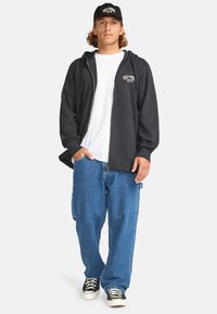 Zwarte ritssluiting hoodie met voorzakken, wit T-shirt, losse blauwe jeans en zwarte pet met geborduurd logo, gecombineerd met zwarte sneakers.