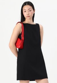 Schwarzes ärmelloses Kleid mit quadratischem Ausschnitt, begleitet von einer strukturierten roten Handtasche mit einem kontrastierenden Träger. Glatte Textur, modernes Design.