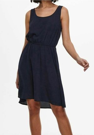 Kvinde iført en ærmeløs, knælange sort kjole med indsnøring i taljen mod en hvid baggrund.