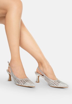 Gambe femminili incrociate che indossano scarpe slingback intrecciate argento con tacco kitten su sfondo chiaro.