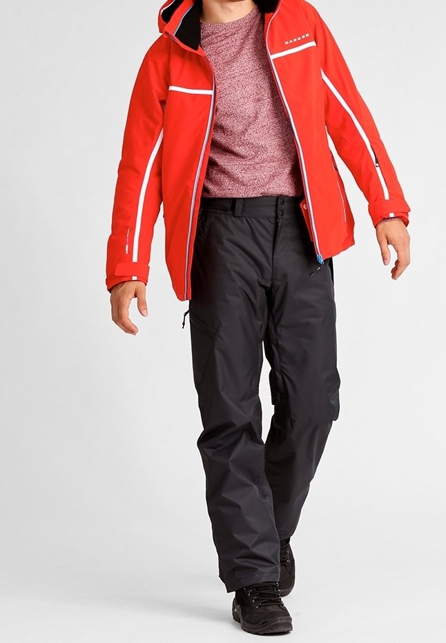 Homme portant une veste à capuche rouge, un pull bordeaux, un pantalon noir et des chaussures noires marchant devant un fond clair uni.