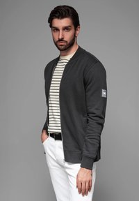 Chaqueta bomber gris oscuro con cremallera frontal y puños de canalé, superpuesta sobre una camiseta de rayas blancas y negras, combinada con pantalones blancos.