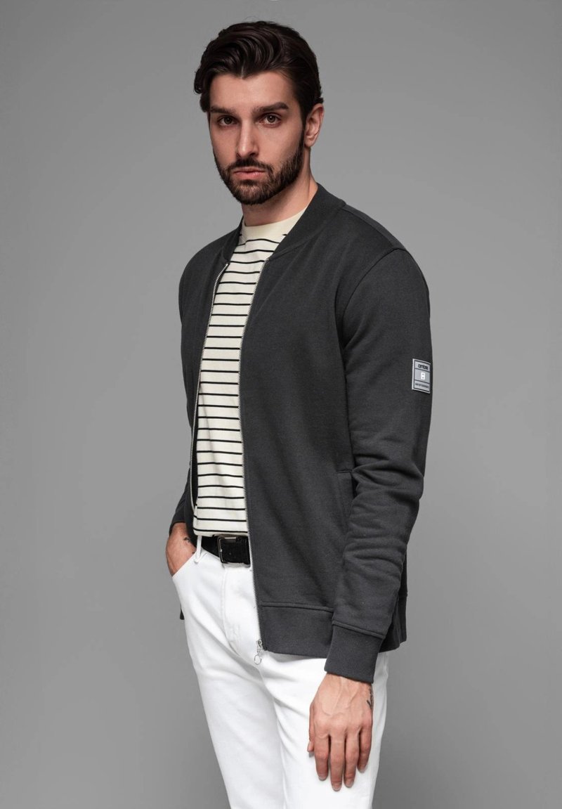 Chaqueta bomber gris oscuro con cremallera frontal y puños de canalé, superpuesta sobre una camiseta de rayas blancas y negras, combinada con pantalones blancos.