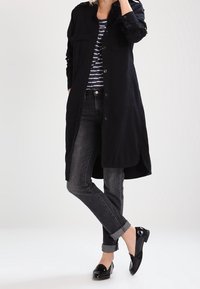 Osoba w czarnym długim płaszczu, w pasiastą koszulę, z podwiniętymi ciemnymi dżinsami i czarnymi błyszczącymi mokasynami, stojąca z krzyżującymi się nogami.