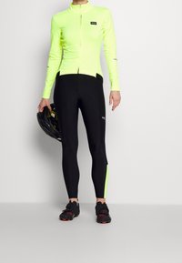 Kirkkaan neon keltainen pitkähihaine paita, jossa on vetoketju, yhdistettynä mustiin legginsseihin, joissa on vihreä sivupaneeli. Malli pitää kädessään mustaa polkupyörä kypärää.