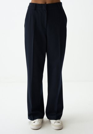 Pantaloni a gamba larga color navy realizzati in tessuto morbido. Presentano una vita alta, pieghe nella parte anteriore e una vestibilità rilassata. Abbinati a sneakers bianche.