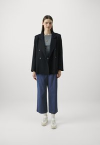 Blazer doppiopetto navy sopra un top grigio, abbinato a pantaloni blu a gamba larga e sneakers bianche. Tessuto morbido e vestibilità rilassata.
