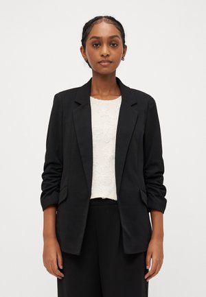 Sorte skreddersydd blazer med en strukturert design, notched revers og trekvart ermer. Brukt over en teksturert hvit topp.