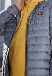 Person iført en grå quiltet jakke over en sennepsgul hættejakke, hånd hævet til halsen, delvist udendørs bybaggrund synlig.