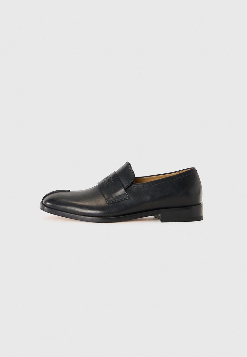 Mocassins en cuir noir avec une texture lisse, un bout rond et une sangle proéminente à l'avant ; présentent un design minimaliste.