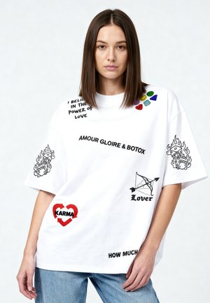 Femme aux cheveux bruns raides porte un T-shirt blanc oversize avec du texte, des symboles comme des dés en feu, un cœur avec "KARMA" et un nœud intitulé "Lover."