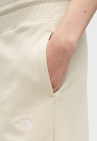 Χέρι στην τσέπη μπεζ φόρμας North Face με ορατό κεντημένο λογότυπο κοντά στη ραφή της τσέπης.