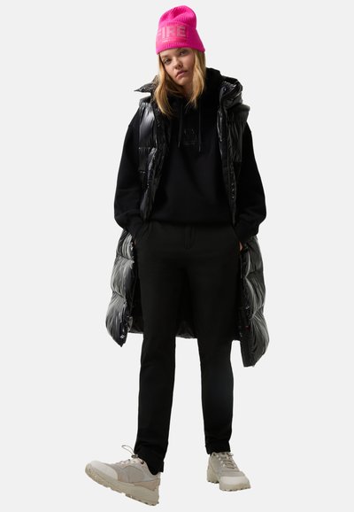 Junge Frau, die eine leuchtend rosa Strickmütze, einen schwarzen Hoodie, eine glänzende lange Daunenweste, schwarze Hosen und beige Sneaker trägt, steht mit den Händen in den Taschen.