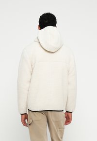 Höyryävä valkoinen fleece-takki, jossa on huppu, ja se on varustettu teksturoidulla pinnalla sekä yksivärisellä kuviolla. Mustat reunat hihansuissa ja helma.