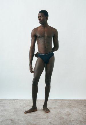 Atleetlik mees seisab paljajalu vaibal, kandes tumedat Tommy Hilfigeri pesu, vaadates vasakule tavalise heleda tausta vastu.