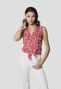 Top sin mangas rojo y blanco con un patrón de hojas, con un detalle de lazo en la parte frontal. Combinado con jeans blancos ajustados.