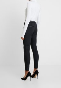 Schwarze, maßgeschneiderte Hose mit schmaler Passform, mit einer glatten Textur und zwei Gesäßtaschen. Kombiniert mit einem figurbetonten weißen Langarmoberteil und schwarzen Absätzen.