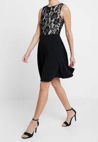 Robe noire avec un corsage en dentelle cintré et une jupe évasée. Le tissu est lisse, et la robe a un col rond et une coupe sans manches.