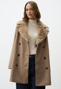 Cappotto in lana beige con colletto in pelliccia sintetica, design doppiopetto, grandi bottoni e vestibilità rilassata, abbinato a un dolcevita chiaro e jeans scuri.