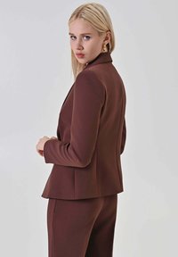 Blazer marrone su misura con una forma aderente, spalle strutturate e una texture liscia, abbinato a pantaloni coordinati dello stesso colore.