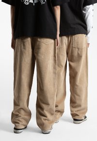 Beige kordfløyelsbukser med bred benpassform, med fremtredende vertikale ribber og baklommer, kombinert med svarte joggesko.