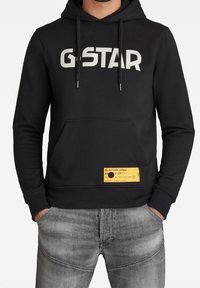 Sudadera negra con el logo "G-STAR" en blanco, bolsillo frontal, etiqueta amarilla en el dobladillo y capucha con cordón. Combinada con vaqueros grises.