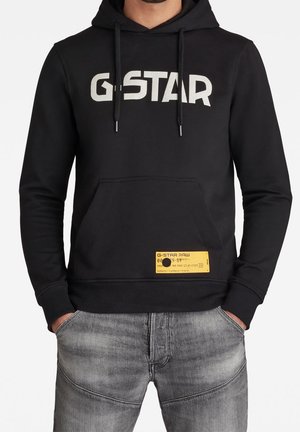 Sudadera negra con el logo "G-STAR" en blanco, bolsillo frontal, etiqueta amarilla en el dobladillo y capucha con cordón. Combinada con vaqueros grises.