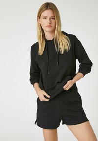 Sort hættejakke med snore, der har lange ærmer og en frontlomme. Sammen med sorte shorts, begge lavet af blødt, glat stof.
