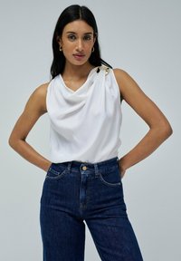 Top sin mangas blanco con tela drapeada y un acento de hardware dorado en el hombro, combinado con jeans de mezclilla oscuros de tiro alto.