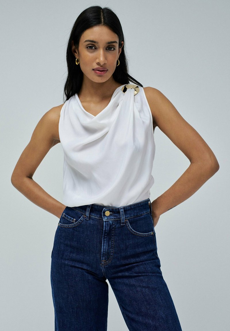 Top sin mangas blanco con tela drapeada y un acento de hardware dorado en el hombro, combinado con jeans de mezclilla oscuros de tiro alto.